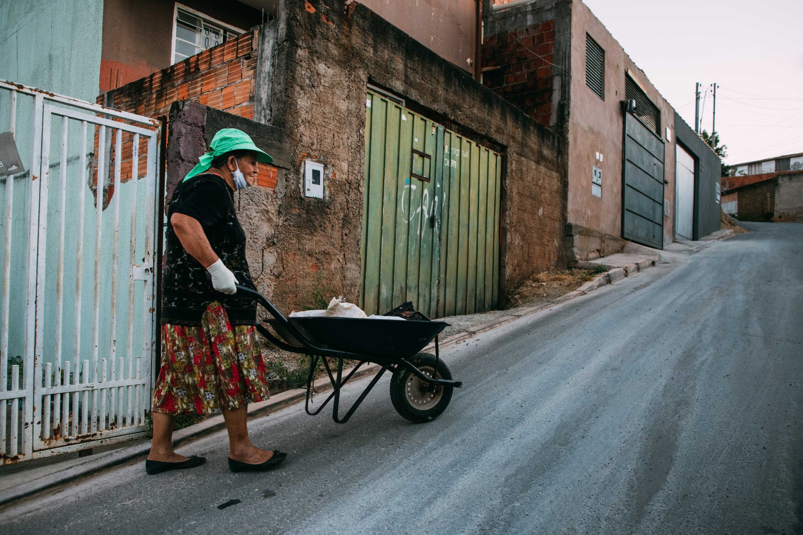 Uma pessoa usando chapéu e máscara empurra um carrinho de mão carregado de sacolas recicláveis em uma rua íngreme em uma área urbana.